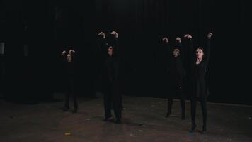 Five confident actors at a theater university dressed in black clothes simultaneously relax and tense individual parts of the body making a synchronized movement during acting training in a theater video