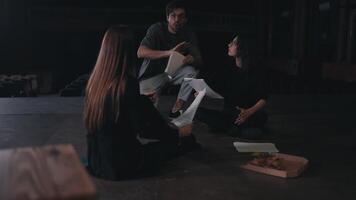 Two female actresses in black suits sit on stage and communicate holding in their hands the text of their characters and the script of the play while their director runs up to them and points out how video
