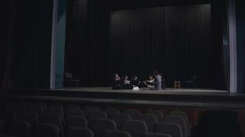 A group of young actors in black uniforms sit on stage together with their stage director communicate and analyze the script while preparing for a performance in a theater with a black curtain video