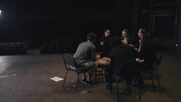 A group of happy actors in black uniforms sit around an impromptu pizza and chat during a break and preparation for a performance on stage in a theater video