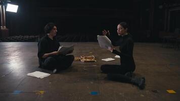 A couple of confident actors a brunette girl in a black uniform and a guy with a kit of papers and a script play their roles and communicate about the script while sitting on stage in an empty theater video