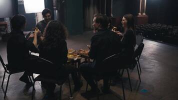 Close-up over the shoulder of confident theater actors in black suits sitting on chairs around and eating pizza during a break and preparation for the performance on stage in the theater video