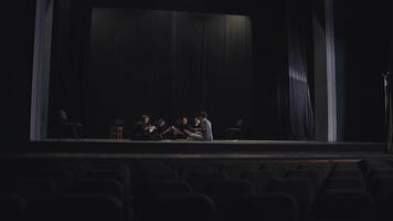 View from afar a group of confident young actors in black suits sit on stage in a theater and have lunch during a break during a rehearsal in a large theater with a black curtain video
