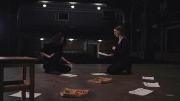 A confident girl with brown hair and a black suit holds sheets of paper with a script in her hands and expressively addresses her fellow actress during her rehearsal for a play on the theater stage video