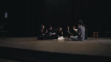 A group of young confident actors in black uniforms are talking with the director about the script for their next play and are sitting on stage in the theater video