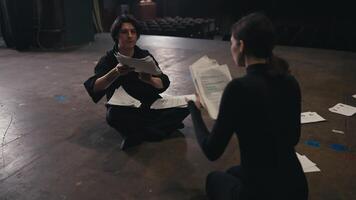 Two confident actors a brunette girl and a man in a black suit are sitting on stage sorting through sheets of paper with a script talking about their role and rehearsals for the play video