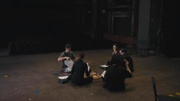 A group of confident actors along with their stage director are sitting on stage having lunch and discussing scripts for their next performance during a rehearsal and work in a theater on stage video