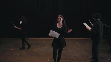 A confident young girl with glowing hair color in a black suit comes closer and reads out her text from a piece of paper while playing her role in the theater impression of emotions on stage during a video