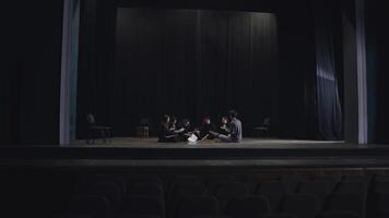 A group of confident young actors in black suits together with their stage director are sitting on stage and preparing for their next performance sorting through sheets of script on stage video