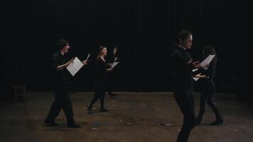 A group of confident actors in black suits walk around the stage and read their words from sheets of paper during their rehearsal and preparation for a performance on stage in a large theater with video
