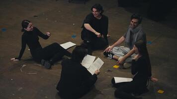 A group of confident young actors in black suits are sitting on stage together with their director holding sheets of paper in their hands with the script of the play on stage in the theater video