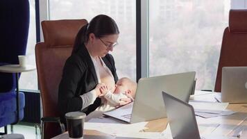 A confident brunette girl in round glasses in a business uniform sits at a table in front of a laptop in the office and breastfeeds her baby without being distracted from work in a modern office video