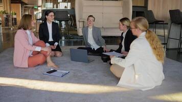 A group of girls in business suits sit on a gray carpet in the office and communicate with each other during a break at work in a modern office video