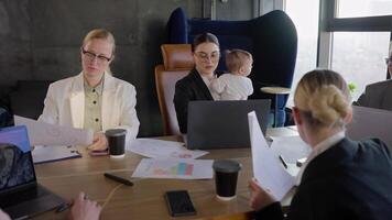 A group of businesswomen in business suits are having a meeting while a colleague is holding a small infant child in her arms while sitting at a table in front of a laptop in a modern office video
