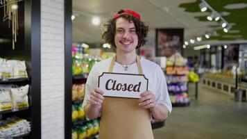 A smiling guy with curly hair and yellow front stands in a supermarket and holds a sign in front of his chest that says Closed video