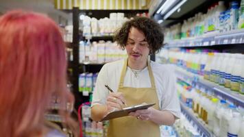 Over the shoulder a happy guy with curly hair holds a tablet in his hands and does an inventory while communicating with customers in the dairy department of a supermarket video