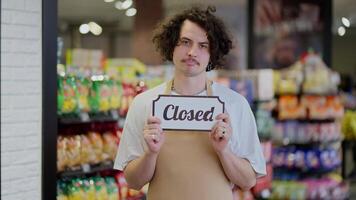 Portrait of a sad brunette guy in an apron who holds in his hands a sign with the inscription closed in a supermarket video