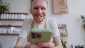 Close up of a happy blond guy with a beard in a white T-shirt looking at social networks using a green phone leaning on the counter in a cafe video