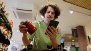 Confident guy with curly brunette hair wearing a green T-shirt scrolling through social networks while shopping in a supermarket with a cart video