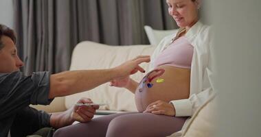 Side view of a happy man and his pregnant wife painting on their belly with bright colors while dreaming about a future child and expecting a child at home video