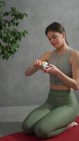 Vertical of a happy brunette girl in gray top green pants and a summer sports uniform lights a candle before her exercise session on a red sports carpet at home in a modern apartment video