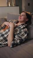 Vertical of a happy and joyful brunette girl in a checkered shirt and an orange top looks at the social network using a white smartphone and communicates online in a modern apartment lying on video