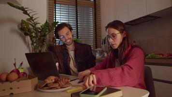 A confident and hardworking brunette girl with glasses in a red sweater studies foreign languages and writes notes in her notebook together with a young brunette male teacher in a modern apartment in video