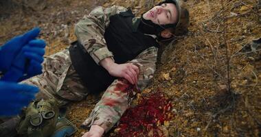 A confident male medic in a dark green army uniform bandages the wounded hand of a soldier in a camouflage uniform with a tourniquet and provides him with first aid during combat operations in the video