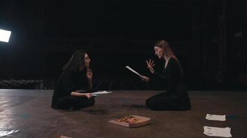 Confident two female actresses in black suits sit on stage holding papers with a script in their hands and expressively expressing their ideas and acting out roles during a rehearsal for a play in an video