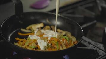 Close-up a cook adds cream to fried vegetables in a hot frying pan and stirs the contents while preparing a dish on a gas burner in a restaurant video
