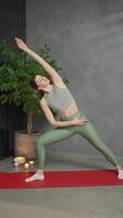Vertical of a confident athletic brunette girl in a gray top and green sweatpants stands on the red mat for exercising near burnt candles and a large home plant during her exercise and video