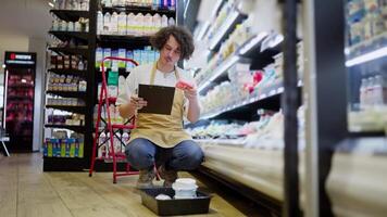 A guy with curly black hair takes an item from the counter and checks it against the inventory and then writes off the expired item in the dairy section of the supermarket video