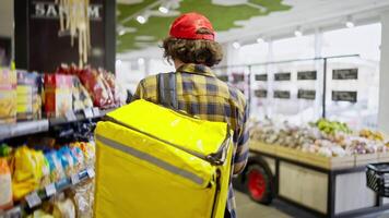 Rear view of a confident brunette guy with curly hair in a checkered shirt with a yellow bag a food delivery man walks near the counter in a supermarket and selects the necessary products video