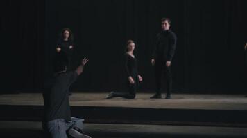 Rear view of a confident male director with papers in his hands waving his arms and gesticulating showing the actors how to play correctly during a rehearsal of actors in black suits on stage in the video