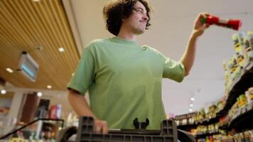 Bottom view of a confident brunette guy with curly hair in a Green T-shirt carries a cart in a supermarket and puts the things he needs from it during his shopping video