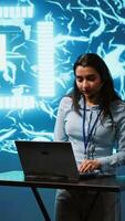 Vertical Technician in high tech workspace managing call center tasks, speaking into headset while doing monitoring with laptop. AI company employee talking with clients through headphones mic, camera A video