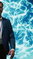 Vertical Upbeat engineer in startup doing advancements in AI field. Portrait of happy man standing with arms crossed in front of cutting edge display featuring AI and neural network graphics, camera A video