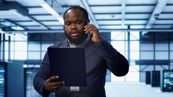IT professional looking over data center checkup checklist paperwork, talking on phone in server farm. African american man surrounded by server rigs, chatting during telephone call, camera A video