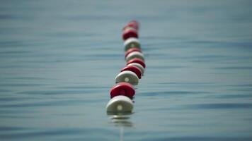 Orange and white safety buoys on a rope floating in the sea on a sunny day, close up. Fencing of the swimming area on the beach. Vacation, travel and holiday concept. Slow motion video
