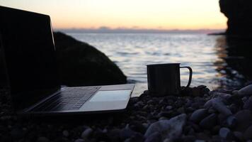 Silhouette of laptop and cup on the beach over sea sunset. Sparkling sea water on background. Freelancer, blogger working on notebook on the beach. video