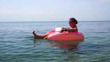 Woman freelancer works on laptop swimming in sea on pink inflatable ring. Happy tourist in sunglasses floating on inflatable donut and working on laptop computer in calm ocean. Remote working anywhere video