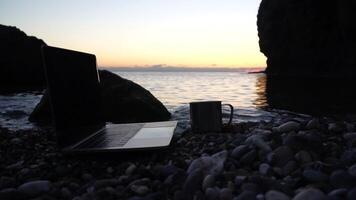Silhouette of laptop and cup on the beach over sea sunset. Sparkling sea water on background. Freelancer, blogger working on notebook on the beach. video