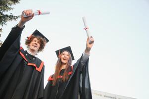 Portrait of happy graduates. Two friends in graduation caps and gowns standing outside university building with other students in background, holding diploma scrolls, and smiling photo