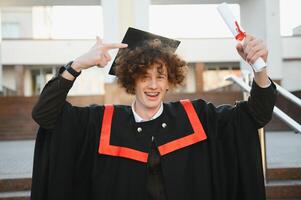 Graduation from university. Young smiling boy university graduate in traditional bonet and mantle standing and holding diploma in hand over university building background photo