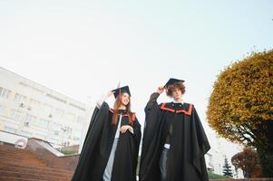 Portrait of happy graduates. Two friends in graduation caps and gowns standing outside university building with other students in background, holding diploma scrolls, and smiling photo