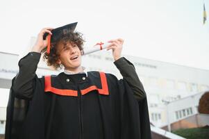 Happy smiling university graduate in mantle holding diploma in raised hand and expressing happiness over university building at background. Successful graduating from university or college photo
