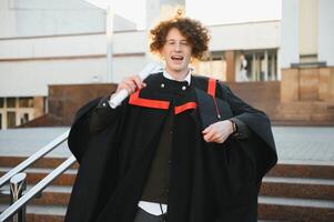 Graduation from university. Young smiling boy university graduate in traditional bonet and mantle standing and holding diploma in hand over university building background photo