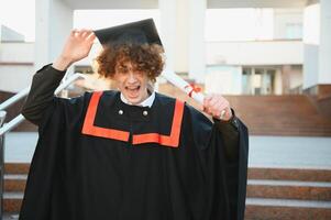 Graduation from university. Young smiling boy university graduate in traditional bonet and mantle standing and holding diploma in hand over university building background photo