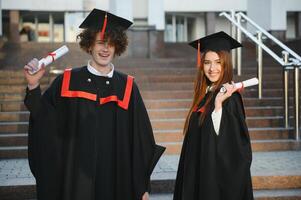 Portrait of happy graduates. Two friends in graduation caps and gowns standing outside university building with other students in background, holding diploma scrolls, and smiling photo