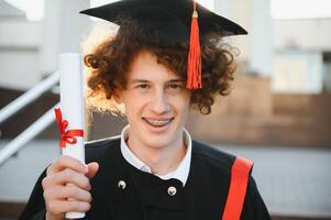 Graduation from university. Young smiling boy university graduate in traditional bonet and mantle standing and holding diploma in hand over university building background photo